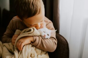 A young boy hugging and kissing a white cat wrapped in a beige blanket